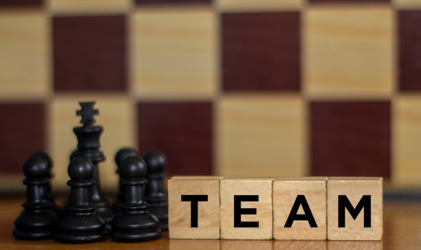 Wooden blocks with the word 'TEAM' are placed next to black chess pieces, including pawns and a king. The background features a blurred chessboard pattern with alternating light and dark squares.