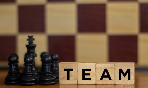 Wooden blocks with the word 'TEAM' are placed next to black chess pieces, including pawns and a king. The background features a blurred chessboard pattern with alternating light and dark squares.