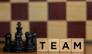 Wooden blocks with the word 'TEAM' are placed next to black chess pieces, including pawns and a king. The background features a blurred chessboard pattern with alternating light and dark squares.
