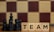 Wooden blocks with the word 'TEAM' are placed next to black chess pieces, including pawns and a king. The background features a blurred chessboard pattern with alternating light and dark squares.