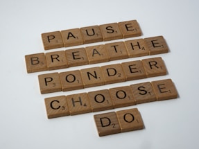 Wooden Scrabble tiles spelling out the words: PAUSE, BREATHE, PONDER, CHOOSE, DO. The tiles are arranged in a grid format with each word on its own line, suggesting a sequence or process. The surface is light, and the tiles have a natural wooden texture.