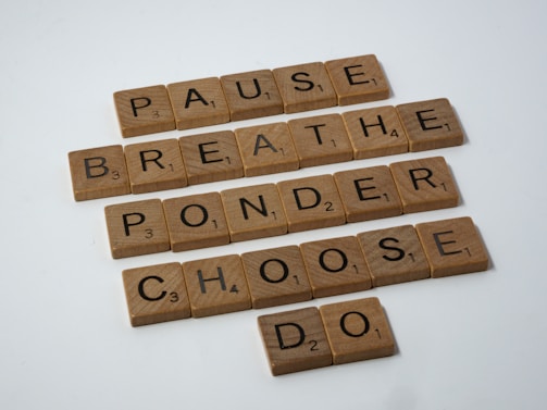 Wooden Scrabble tiles spelling out the words: PAUSE, BREATHE, PONDER, CHOOSE, DO. The tiles are arranged in a grid format with each word on its own line, suggesting a sequence or process. The surface is light, and the tiles have a natural wooden texture.