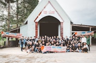 A large group of people are gathered in front of a building with a triangular roof and a sign that reads 'TIN LÀNH'. They appear to be participating in a community event, posing for a group photo. Several banners with the text 'GIEO NANG' are held by some group members. The setting appears to be outdoors with trees surrounding the building.