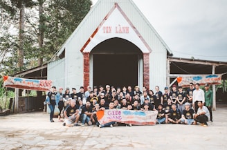 A large group of people are gathered in front of a building with a triangular roof and a sign that reads 'TIN LÀNH'. They appear to be participating in a community event, posing for a group photo. Several banners with the text 'GIEO NANG' are held by some group members. The setting appears to be outdoors with trees surrounding the building.