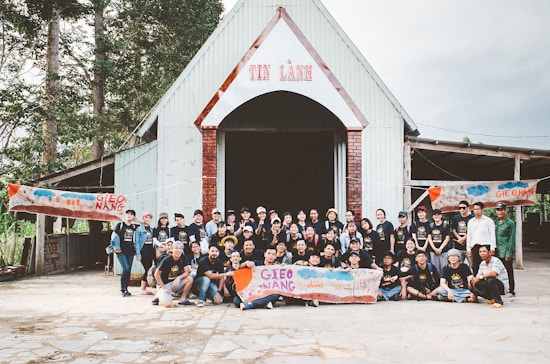 A large group of people are gathered in front of a building with a triangular roof and a sign that reads 'TIN L&Agrave;NH'. They appear to be participating in a community event, posing for a group photo. Several banners with the text 'GIEO NANG' are held by some group members. The setting appears to be outdoors with trees surrounding the building.