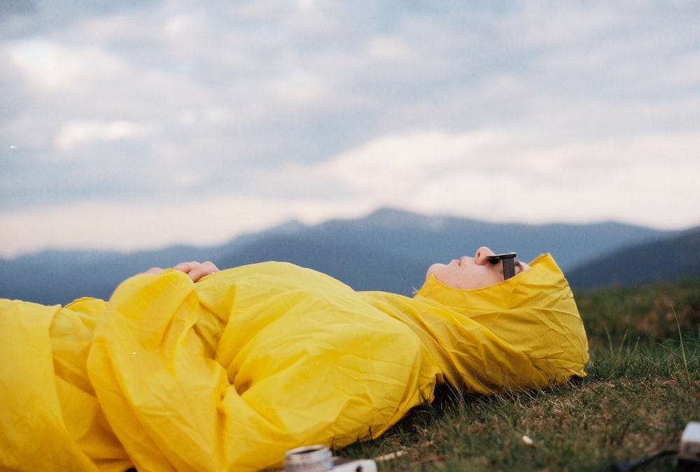昼間、白い曇り空の下、緑の芝生に横たわる黄色いパーカーを着た男