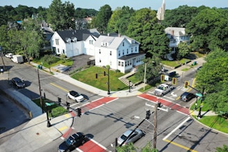 An aerial view of a residential neighborhood featuring several large houses surrounded by greenery. There are cars at a four-way stop intersection with visible traffic lights. The streets are lined with trees and the area appears calm and well-maintained.