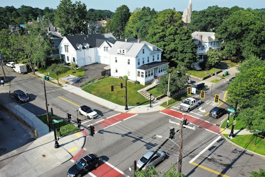 An aerial view of a residential neighborhood featuring several large houses surrounded by greenery. There are cars at a four-way stop intersection with visible traffic lights. The streets are lined with trees and the area appears calm and well-maintained.