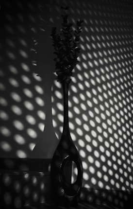A tall, slender vase with a circular opening sits on a surface, casting intricate shadow patterns of light and dark circles on the wall behind it. The scene is in monochrome, highlighting the contrast between the light and shadows.