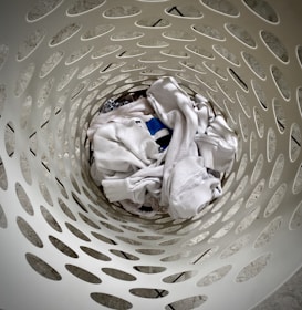 Looking down into a white plastic laundry basket with a perforated pattern, several pieces of white and light-colored clothing are crumpled inside.