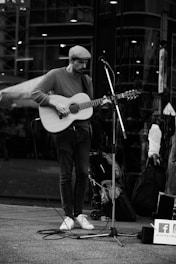 A street musician plays the guitar near a microphone stand in an urban setting. The musician wears a flat cap, sweater, jeans, and sneakers. The background shows reflections of the city architecture on glass. A suitcase and bags are placed nearby, hinting at a traveling performer.