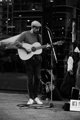 A street musician plays the guitar near a microphone stand in an urban setting. The musician wears a flat cap, sweater, jeans, and sneakers. The background shows reflections of the city architecture on glass. A suitcase and bags are placed nearby, hinting at a traveling performer.