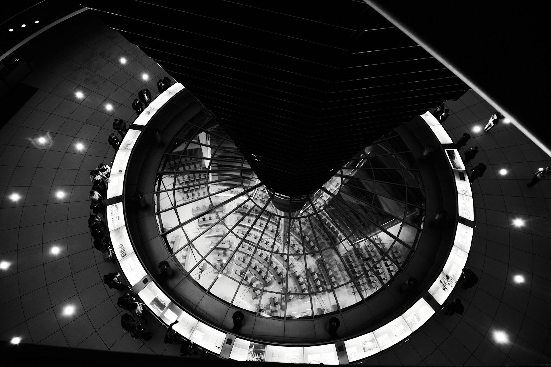 A black and white photograph captures an architectural interior featuring a circular glass structure at the center. The scene is viewed from above, showcasing a spiral or conical element surrounded by a ring of illuminated panels. Several people are positioned around the outer edge, standing or walking across the patterned floor.