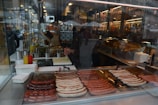 A street food stall with a variety of sausages cooking on a grill inside a glass-enclosed space. Mustard and ketchup bottles are to the side, with buns stacked in the background. Reflections of the street can be seen in the glass.