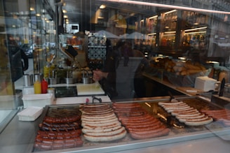 A street food stall with a variety of sausages cooking on a grill inside a glass-enclosed space. Mustard and ketchup bottles are to the side, with buns stacked in the background. Reflections of the street can be seen in the glass.