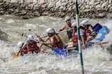 A group of people wearing helmets and life jackets are navigating a raft through turbulent whitewater rapids, surrounded by rocks along the riverbank. They appear to be focused and actively rowing with paddles.
