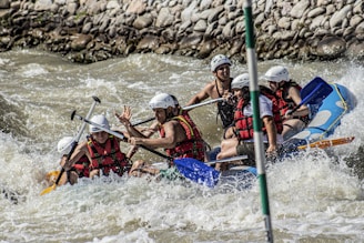 A group of people wearing helmets and life jackets are navigating a raft through turbulent whitewater rapids, surrounded by rocks along the riverbank. They appear to be focused and actively rowing with paddles.