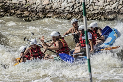 A group of people wearing helmets and life jackets are navigating a raft through turbulent whitewater rapids, surrounded by rocks along the riverbank. They appear to be focused and actively rowing with paddles.