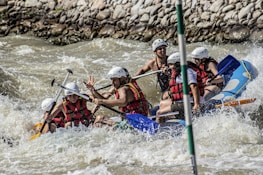 A group of people wearing helmets and life jackets are navigating a raft through turbulent whitewater rapids, surrounded by rocks along the riverbank. They appear to be focused and actively rowing with paddles.