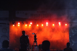 A scene capturing a silhouette of a cameraman with a tripod in front of a brightly lit stage. The stage is illuminated by multiple orange and red lights, producing a foggy, atmospheric effect. The space appears to be a concert or performance venue with additional figures visible.