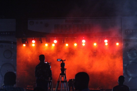 A scene capturing a silhouette of a cameraman with a tripod in front of a brightly lit stage. The stage is illuminated by multiple orange and red lights, producing a foggy, atmospheric effect. The space appears to be a concert or performance venue with additional figures visible.