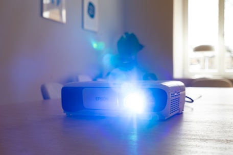 A projector is placed on a wooden table, emitting a bright blue light. The background features a softly lit room with a window that allows natural light to filter in. Minimalistic furniture is visible, including chairs and wall art.