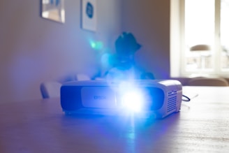 A projector is placed on a wooden table, emitting a bright blue light. The background features a softly lit room with a window that allows natural light to filter in. Minimalistic furniture is visible, including chairs and wall art.