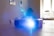 A projector is placed on a wooden table, emitting a bright blue light. The background features a softly lit room with a window that allows natural light to filter in. Minimalistic furniture is visible, including chairs and wall art.