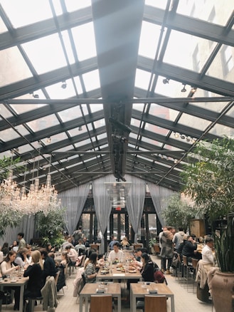 A spacious restaurant featuring a glass ceiling with natural light pouring in. Diners are seated at wooden tables, enjoying meals in a lively atmosphere. Elegant chandeliers and lush green plants add to the ambiance, creating a modern and inviting environment.
