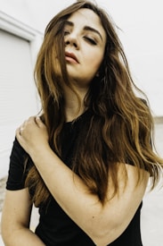 A woman with long brown hair stands against a light background, gently holding her own shoulder. Her eyes are closed and her facial expression appears serene and contemplative. She wears a black top, and her hair partially covers her face.