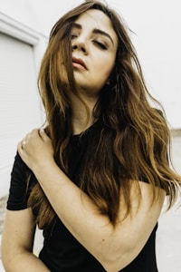 A woman with long brown hair stands against a light background, gently holding her own shoulder. Her eyes are closed and her facial expression appears serene and contemplative. She wears a black top, and her hair partially covers her face.