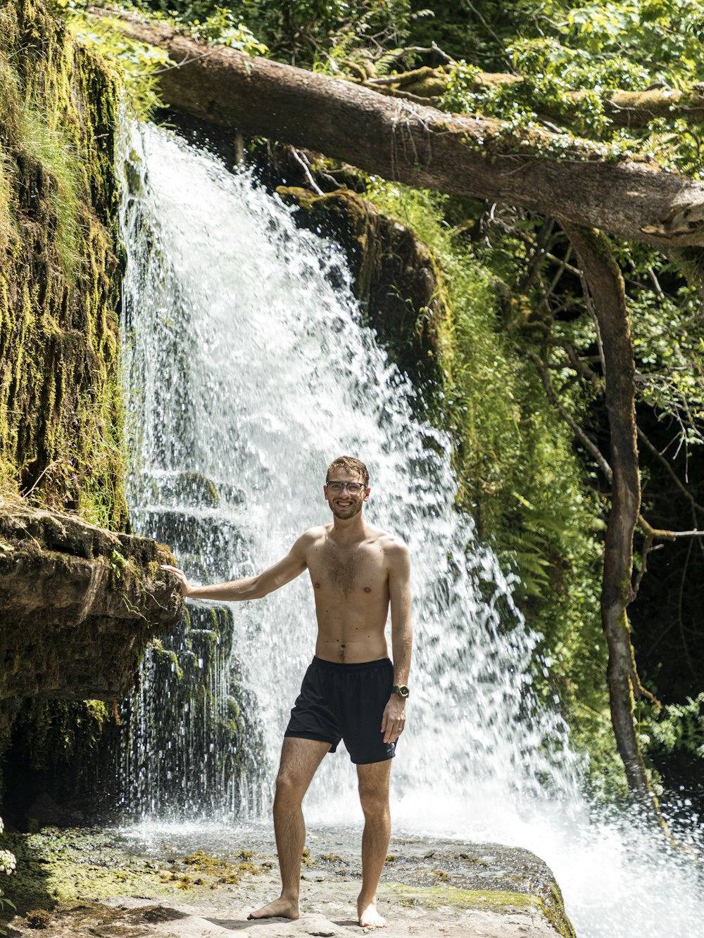 man in blue shorts standing on brown rock near water falls during daytime