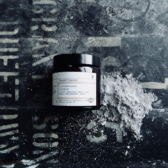 A dark glass jar with a label reading 'Gunpowder Fragrance' sits on a textured surface. Next to the jar is a pile of gray powder, possibly representing the contents or theme of the fragrance.