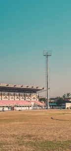 A large outdoor sports stadium with a green field and red spectator stands. A tall floodlight tower stands prominently beside the stands. A few people are walking and congregating near the seating area. The surrounding area includes some palm trees and a few buildings. The sky is clear and blue, suggesting a sunny day.