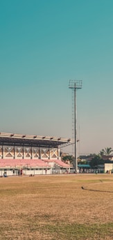 A large outdoor sports stadium with a green field and red spectator stands. A tall floodlight tower stands prominently beside the stands. A few people are walking and congregating near the seating area. The surrounding area includes some palm trees and a few buildings. The sky is clear and blue, suggesting a sunny day.