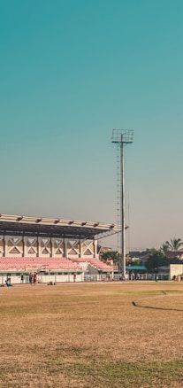 A large outdoor sports stadium with a green field and red spectator stands. A tall floodlight tower stands prominently beside the stands. A few people are walking and congregating near the seating area. The surrounding area includes some palm trees and a few buildings. The sky is clear and blue, suggesting a sunny day.