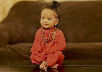 A young child with a cheerful expression is dressed in a red traditional outfit, adorned with beaded necklaces. The child's hair is styled in a topknot, and there is an ornamental mark on the forehead. The background features a dark brown couch.
