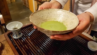 A person is holding a ceramic bowl containing green matcha tea. The bowl is speckled with a rustic texture. To the side, there is a small blue and white tea cup on a wooden surface, possibly a tea tray.