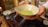 A person is holding a ceramic bowl containing green matcha tea. The bowl is speckled with a rustic texture. To the side, there is a small blue and white tea cup on a wooden surface, possibly a tea tray.