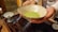 A person is holding a ceramic bowl containing green matcha tea. The bowl is speckled with a rustic texture. To the side, there is a small blue and white tea cup on a wooden surface, possibly a tea tray.