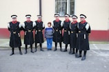 A group of men dressed in ceremonial military uniforms stand in a row alongside a woman in casual clothing. The uniforms are dark with red and gold accents, and the men wear matching hats. The background features a light-colored building with a window.