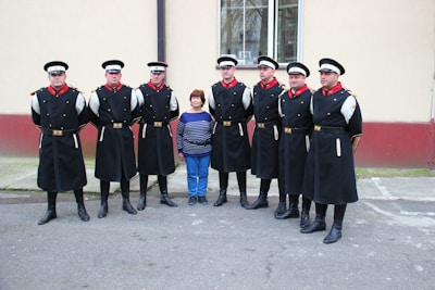 A group of men dressed in ceremonial military uniforms stand in a row alongside a woman in casual clothing. The uniforms are dark with red and gold accents, and the men wear matching hats. The background features a light-colored building with a window.