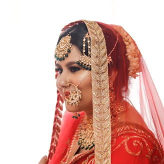 A woman dressed in traditional bridal attire with intricate gold jewelry and a red and gold embroidered veil. Her hair is adorned with floral decorations, and she wears a large nose ring.