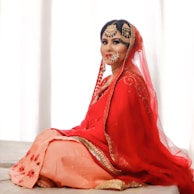 A woman is wearing a traditional red bridal outfit adorned with intricate gold embroidery and jewelry. She is seated gracefully on a light-colored surface, with a serene expression on her face. The veil drapes elegantly over her head and shoulders, enhancing the richness of her attire.