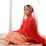 A woman is wearing a traditional red bridal outfit adorned with intricate gold embroidery and jewelry. She is seated gracefully on a light-colored surface, with a serene expression on her face. The veil drapes elegantly over her head and shoulders, enhancing the richness of her attire.