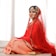 A woman is wearing a traditional red bridal outfit adorned with intricate gold embroidery and jewelry. She is seated gracefully on a light-colored surface, with a serene expression on her face. The veil drapes elegantly over her head and shoulders, enhancing the richness of her attire.