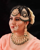 A woman is adorned with elaborate traditional jewelry, including a large maangtika on her forehead, a nose ring with intricate designs, statement earrings, and a heavy necklace with green accents. Her hair is styled with white flowers, adding to the ornate and festive appearance. She is wearing a peach-colored outfit on a dark background.