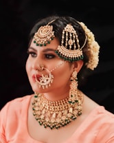 A woman is adorned with elaborate traditional jewelry, including a large maangtika on her forehead, a nose ring with intricate designs, statement earrings, and a heavy necklace with green accents. Her hair is styled with white flowers, adding to the ornate and festive appearance. She is wearing a peach-colored outfit on a dark background.