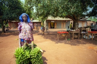 A woman dressed in colorful traditional attire stands in front of a large bunch of green bananas. Behind her, there are stalls displaying tomatoes and other produce under a tree, with several people and bicycles scattered around in a rural setting.