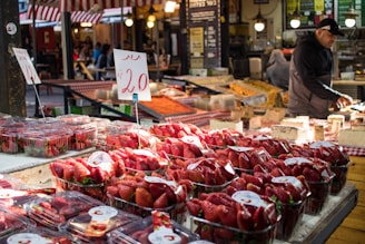 A market stall is filled with containers of fresh strawberries sitting on a wooden counter. Price tags with the number 20 are prominently displayed. A vendor wearing a dark jacket and cap is seen working in the background. The surrounding area includes other produce items and has a bustling atmosphere with various market elements, such as stall signs and lights.
